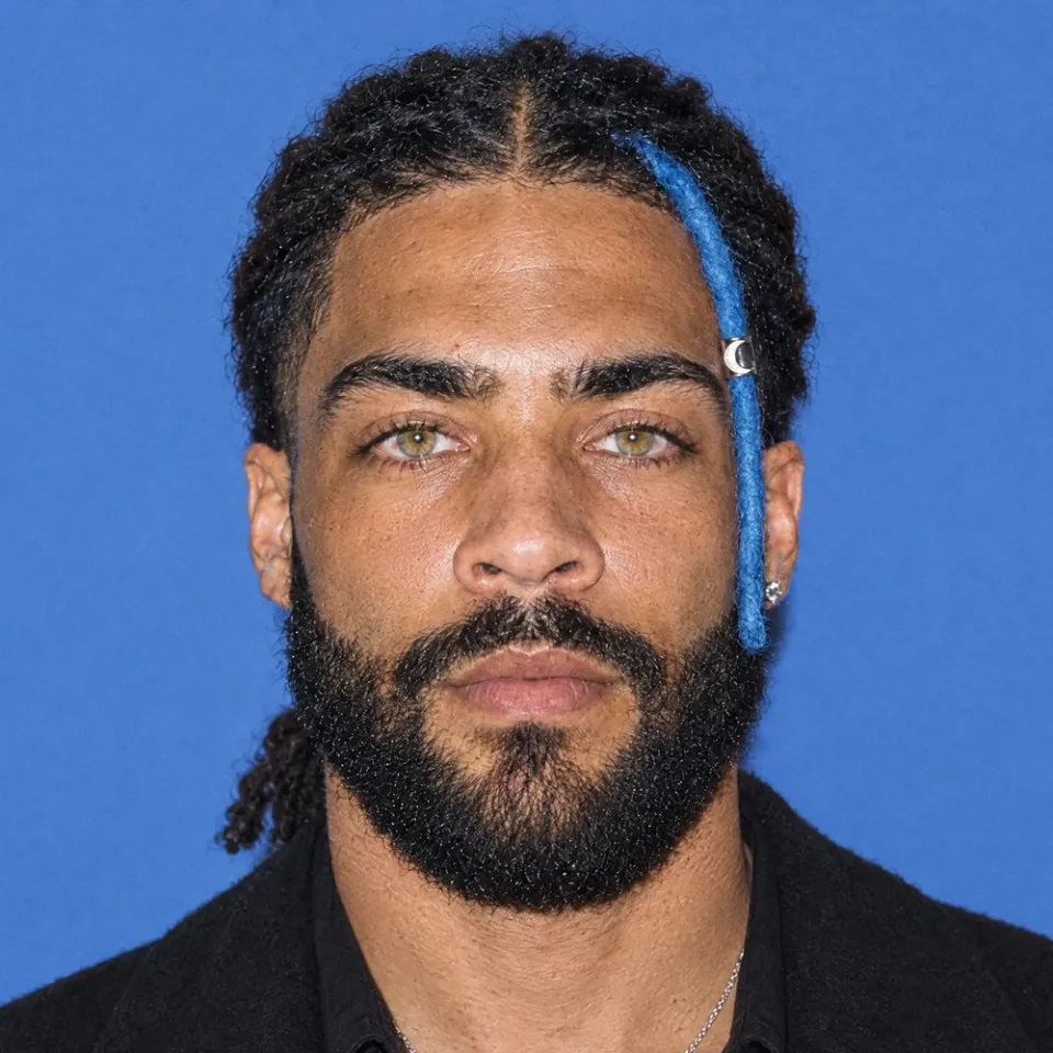 A square close-up on flat blue, straight to camera, the blue loc and cuff hanging at the front of his hair.