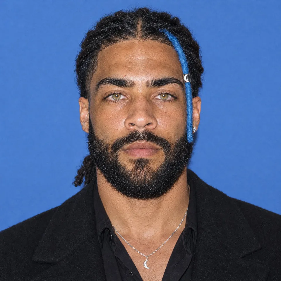 A square portrait on flat blue in a black wool coat over a black shirt, a thin crescent pendant at his chest.