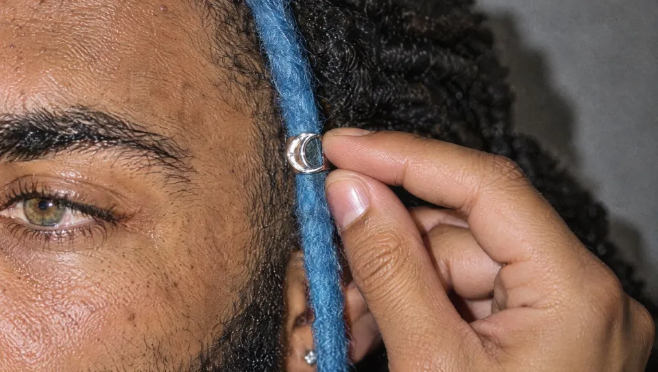 An extreme close-up of his fingers on the silver crescent cuff clamped on the blue loc, beside his hazel-green eye.