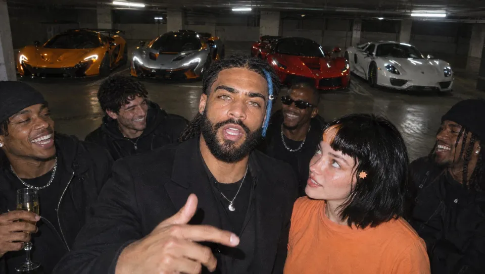 In the garage with friends and Claudia, pointing at the lens, four supercars (orange, silver, red, white) lined up behind.