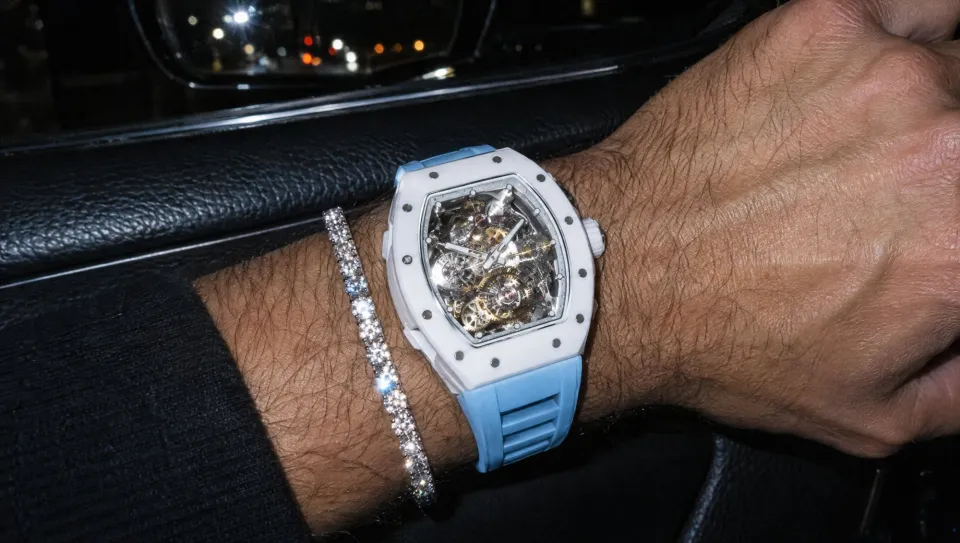 A close-up of his wrist: a white ceramic tonneau skeleton watch on a light-blue strap beside a diamond tennis bracelet.
