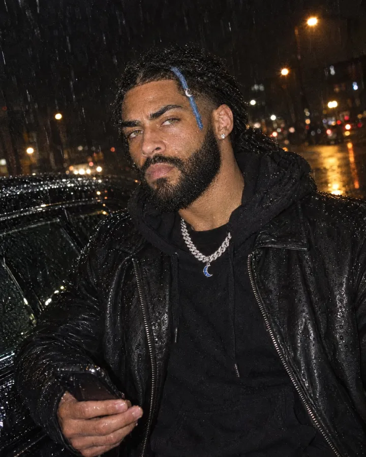 Leaning on a black car in heavy rain, in a wet leather jacket over a hoodie, phone in hand, looking down the lens.