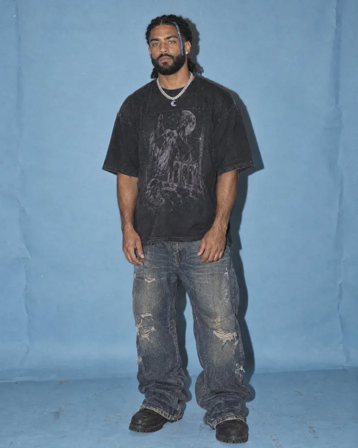 Full length on a blue paper backdrop in an oversized washed-black graphic tee, baggy ripped jeans and black boots.